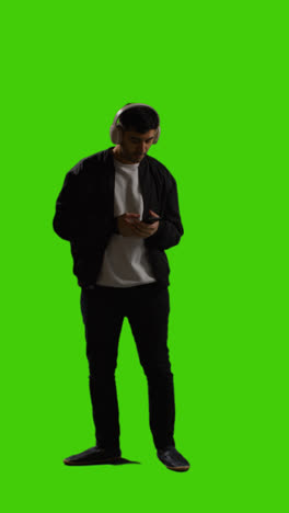 Vertical-Video-Full-Length-Shot-Of-Man-Wearing-Wireless-Headphones-Streaming-Music-From-Mobile-Phone-Against-Green-Screen-1